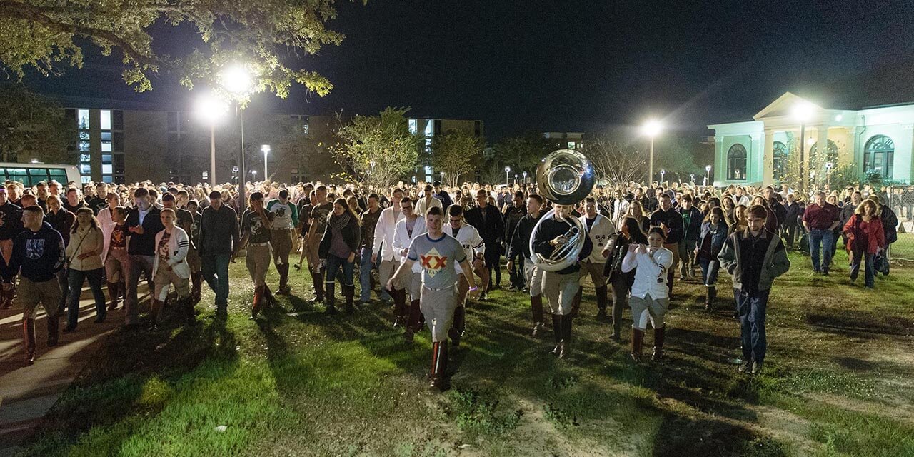 Color photo of Texas A&M students marching at night during Elephant Walk, led by a sousaphone and piccolo.